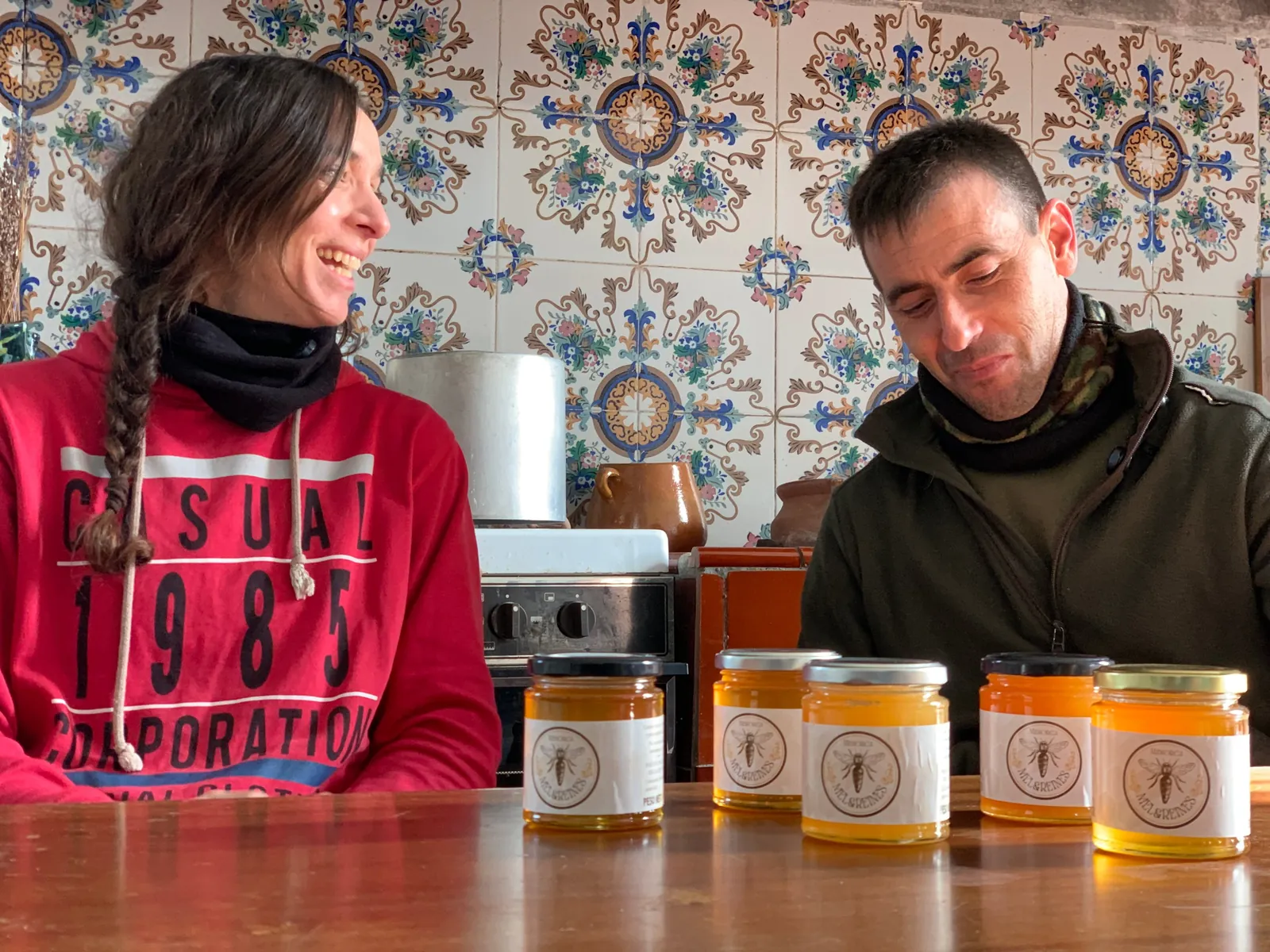 Dos personas sonrientes junto a tarros de miel en una cocina con azulejos